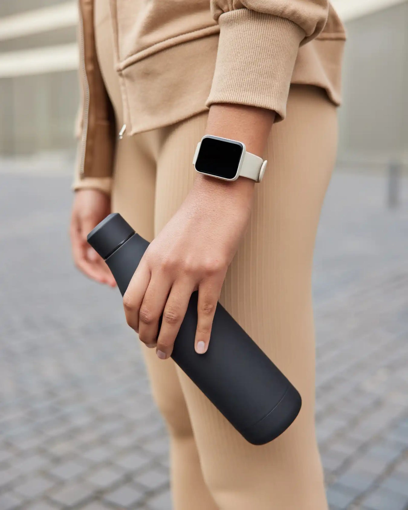 A person wearing a light brown tracksuit and a light-colored smartwatch holding a dark grey water bottle.